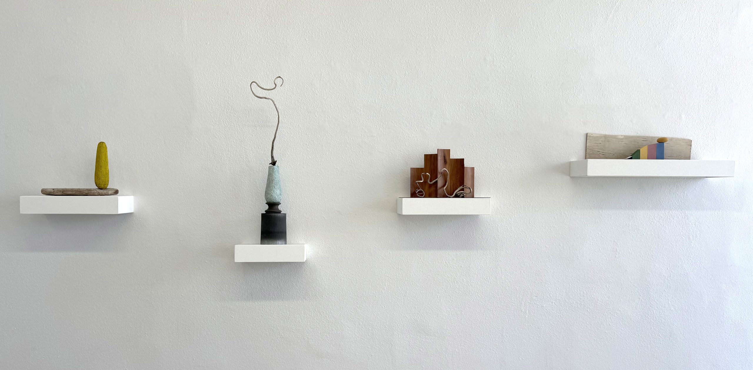 Four curious small sculptures using natural and manmade materials on white shelves against a white wall