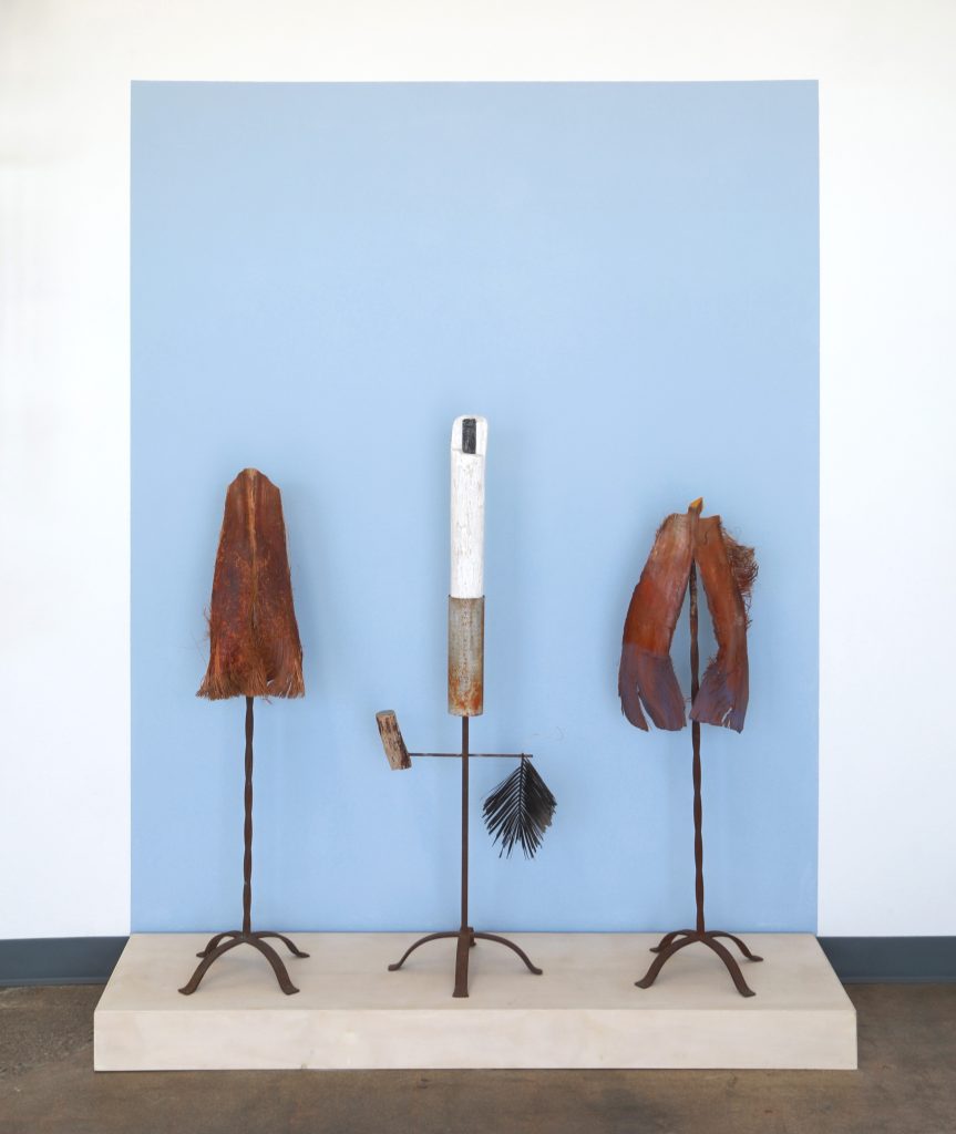 Three metal sculptures on a wooden plinth that support either a palm frond or driftwood, and other materials, suggesting figures